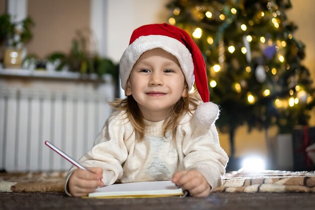 Joli garçon en pull rouge et bonnet rouge écrit une lettre au Père Noël