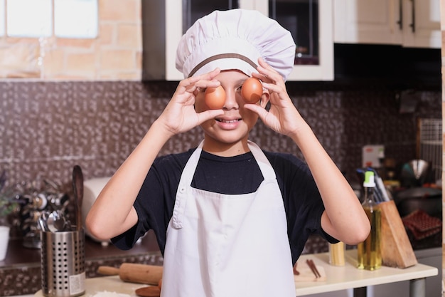 Photo joli garçon utilisant l'uniforme de chef mettant des œufs sur les yeux, se sentant excité à l'idée de cuisiner dans la cuisine