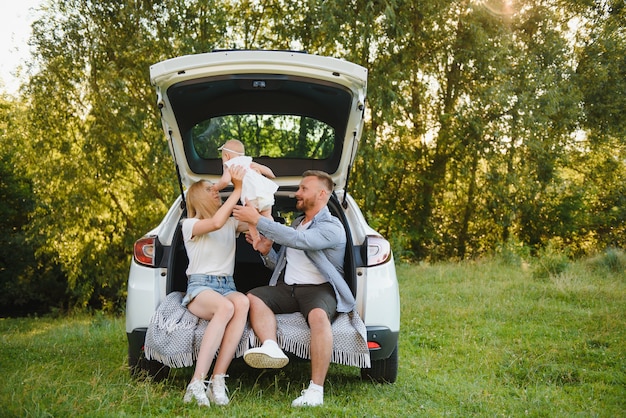 Un joli jeune couple marié et leur fille se reposent dans la nature. La femme et la fille sont assises sur un coffre de voiture ouvert.