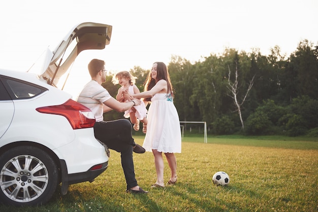 Joli jeune couple marié et leur fille se reposent dans la nature. La mère, le père et la petite fille sont assis sur le coffre de voiture ouvert