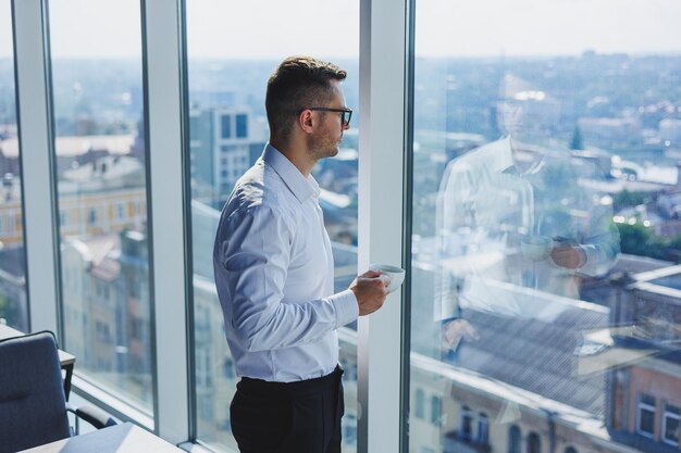 Joli jeune homme d'affaires avec un café à la main dans des verres et une chemise blanche, il est dans un bureau moderne avec de grandes fenêtres Un homme en pantalon classique et une chemise blanche