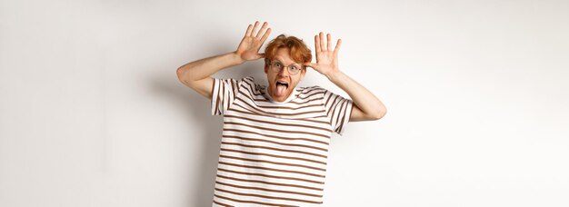 Photo joli jeune homme aux cheveux roux agissant bêtement montrant la langue et se moquant de quelqu'un avec un visage stupide personne debout