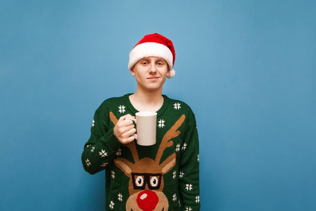 Joli jeune homme en vêtements de noël debout avec une tasse de boisson chaude sur fond bleu