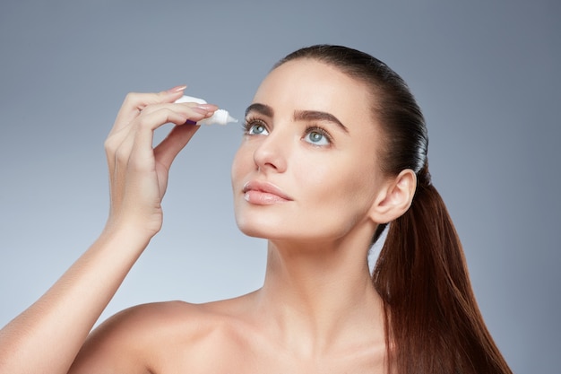 Joli modèle aux cheveux bruns fixés derrière, peau fraîche et propre, grands yeux et épaules nues posant sur fond gris de studio et regardant la caméra, gros plan, à l'aide de gouttes pour les yeux.