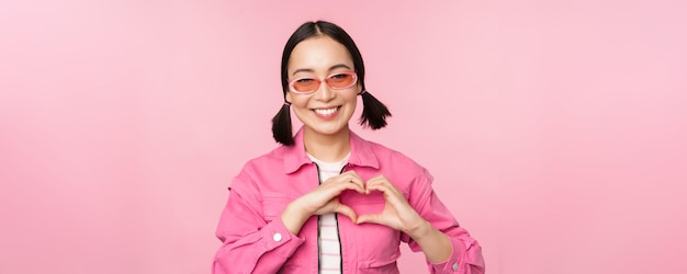 Joli modèle féminin coréen dans des lunettes de soleil à la mode montre un signe de soins cardiaques Je t'aime le geste se dresse sur fond rose