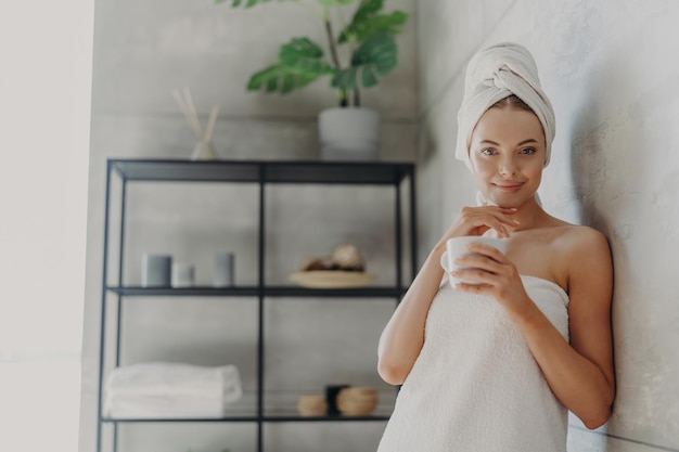 Un joli modèle féminin a une peau parfaite touche la mâchoire boit doucement une boisson chaude se sent rafraîchie et détendue enveloppée dans une serviette de bain pose dans la salle de bain Beauté naturelle et concept de soins de la peau