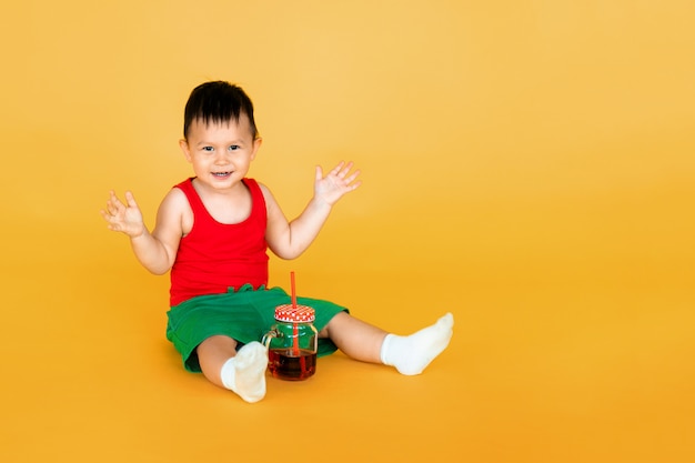 Joli petit bébé garçon en t-shirt rouge et short vert avec une tasse de jus sur jaune