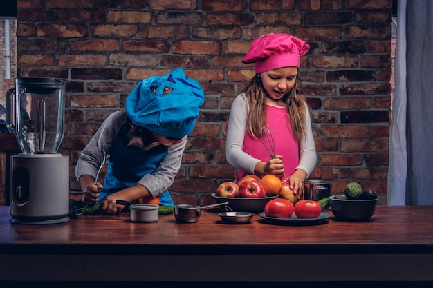 Joli petit couple de cuisiniers. Petit garçon aux cheveux bouclés bruns vêtu d'un uniforme de cuisinier bleu et une belle fille vêtue d'un uniforme de cuisinier rose cuisinant ensemble dans une cuisine contre un mur de briques.