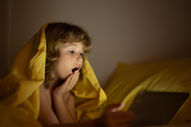 Joli petit enfant couché sous une couverture avec une tablette surpris enfant dans le lit sous la couverture regardant la vidéo sur