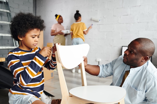 Joli petit garçon aidant son père à assembler la chaise tout en fixant son dos contre la mère et la sœur peignant le mur du salon de couleur blanche