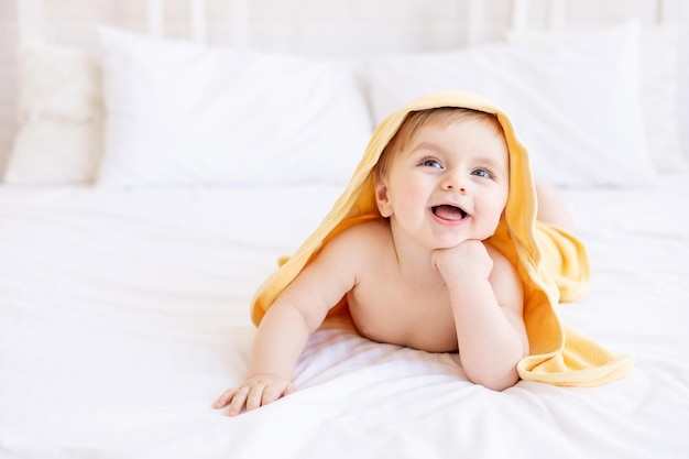 Joli petit garçon blond avec une serviette jaune sur la tête après un bain ou un bain sur un lit blanc à la maison dans une pièce lumineuse sourit le concept de garde d'enfants et d'hygiène