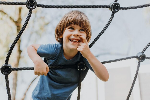 Un joli petit garçon brune dans un T-shirt bleu grimpe sur une toile de corde. l'enfant est engagé dans des sports de plein air. enfant actif en bonne santé. mode de vie. espace pour le texte. Photo de haute qualité