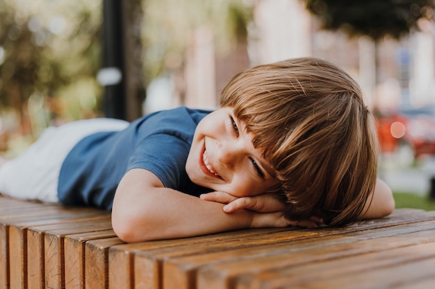 Joli petit garçon brune dans un T-shirt bleu et un short blanc est allongé sur un banc en bois dans le parc. l'enfant prend un bain de soleil et prend un bain d'air à l'extérieur. notion de santé. espace pour le texte. Photo de haute qualité