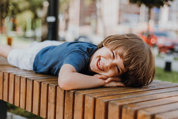 Joli petit garçon brune dans un T-shirt bleu et un short blanc est allongé sur un banc en bois dans le parc. l'enfant prend un bain de soleil et prend un bain d'air à l'extérieur. notion de santé. espace pour le texte. Photo de haute qualité
