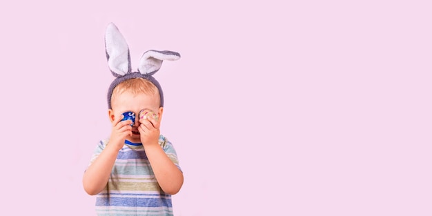 Joli petit garçon dans des oreilles de lapin de lapin sur la tête en fermant les yeux avec des œufs colorés sur fond rose. fête de Pâques