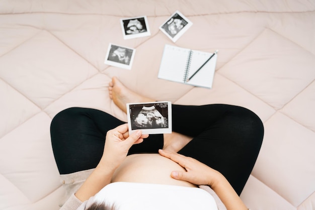 Un joli ventre de femme enceinte et une échographie aux rayons X d'un bébé asiatique jeune femme enceinte se détendre dans la chambre en regardant sa photo d'enfant dans les mains concept de maternité douce troisième trimestre