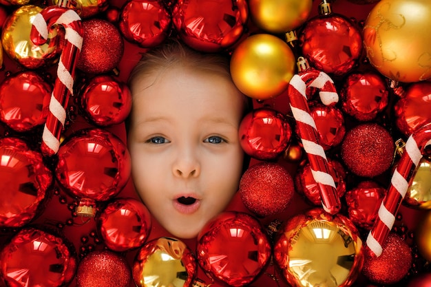 Joli visage enthousiaste étonné d'un enfant dans un tas de boules de Noël rouge et or lumineuses