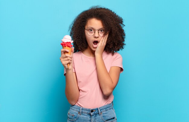 Jolie adolescente afro se sentant choquée et effrayée, l'air terrifiée avec la bouche ouverte et les mains sur les joues. concept de crème glacée sumer