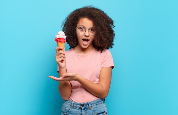 Jolie adolescente afro semblant surprise et choquée, avec la mâchoire tombée tenant un objet avec une main ouverte sur le côté