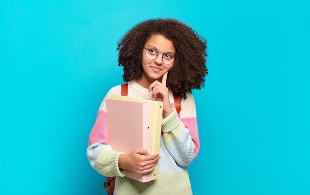 Jolie adolescente afro souriante joyeusement et rêvant ou doutant, regardant sur le côté. concept d'étudiant