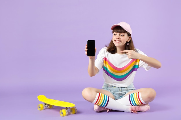 Jolie adolescente assise près de la planche à roulettes, pointant l'index sur le téléphone portable avec un écran vide vide isolé sur fond violet. Émotions sincères des gens, concept de style de vie. Maquette de l'espace de copie.