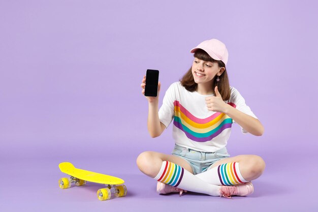 Jolie adolescente assise près de la planche à roulettes tenant un téléphone portable avec un écran vide vide, montrant le pouce vers le haut isolé sur fond violet. Émotions sincères des gens, concept de style de vie. Maquette de l'espace de copie.