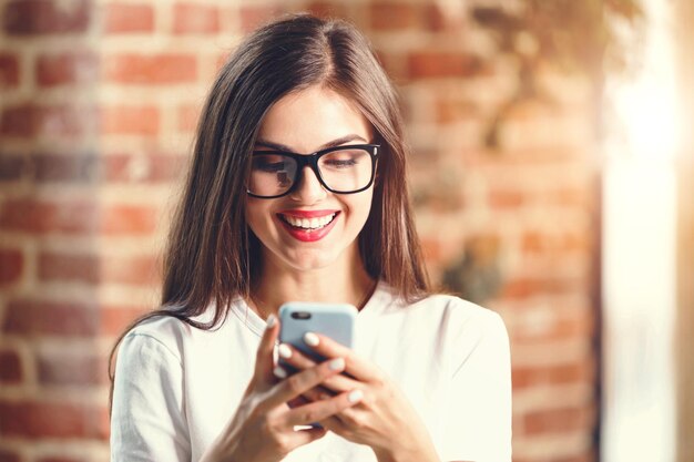 Jolie adolescente aux cheveux longs dans des verres écrire des sms sur le téléphone mobile et souriant dans le bureau moderne du loft