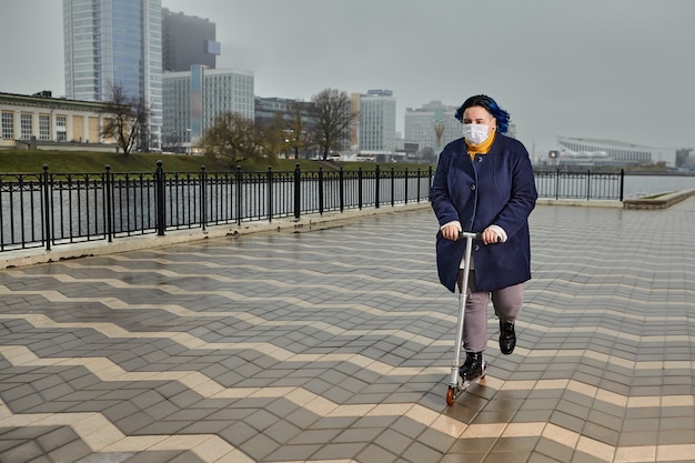 Jolie adolescente blanche aux cheveux bleus fait du scooter le long de la promenade avec masque médical