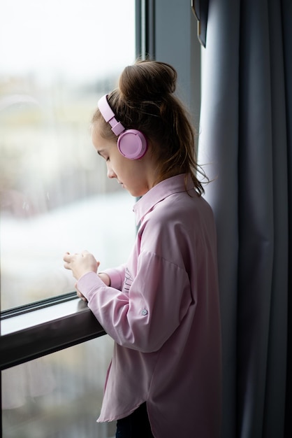 Une jolie adolescente dans une chemise rose et un casque regarde par la fenêtre et rêve