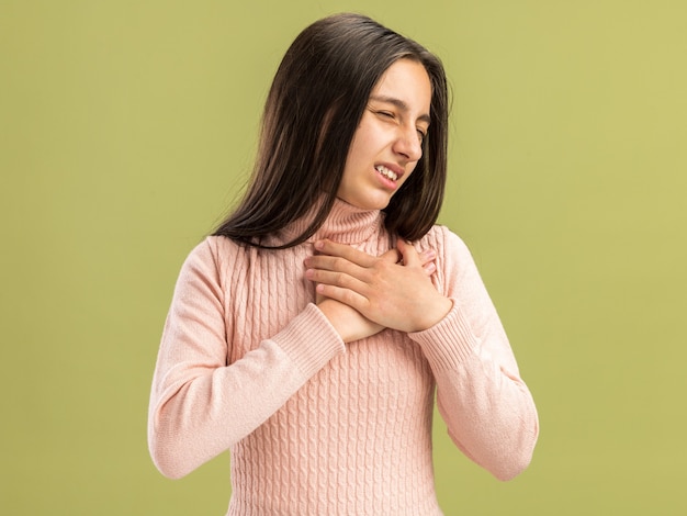 Jolie adolescente douloureuse gardant les mains sur le cœur avec les yeux fermés isolé sur un mur vert olive avec espace pour copie