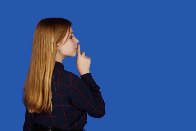 Une jolie adolescente émotionnelle de treize ans dans des verres avec un doigt sur la bouche regarde attentivement, isolée sur fond bleu. Un enfant caucasien a mis le doigt sur les lèvres en signe de silence. Espace de copie