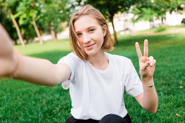 Jolie adolescente faisant selfie dans le parc sur l'herbe et montrant le geste de la paix