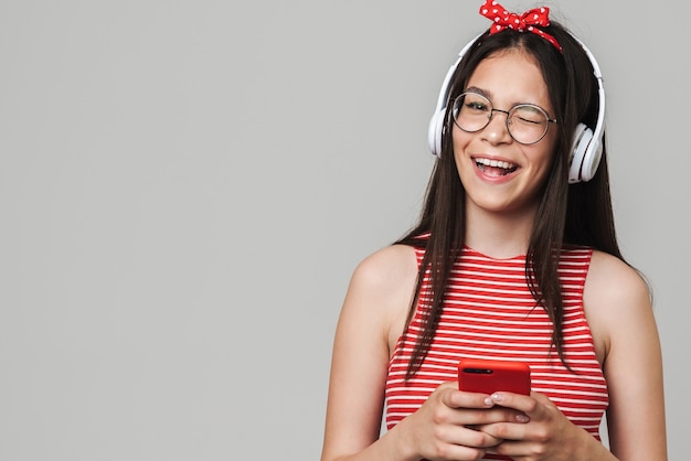 Jolie adolescente joyeuse portant une tenue décontractée, isolée sur un mur gris, écoutant de la musique avec des écouteurs, utilisant un téléphone portable, un clin d'œil