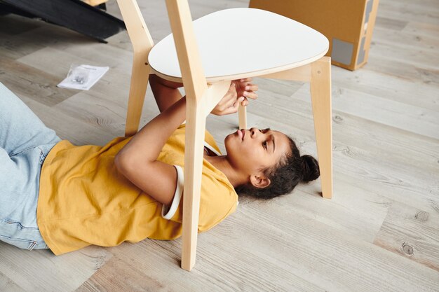 Jolie adolescente métisse en tenue décontractée assemblant une chaise en bois allongée sur le sol du salon et fixant son siège