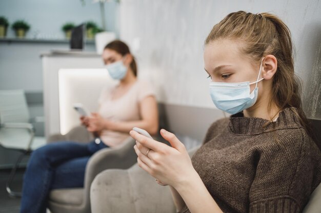 Une jolie adolescente portant un masque facial et utilisant son téléphone portable dans la salle d'attente du cabinet du dentiste.