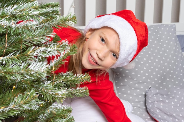 Une jolie adolescente en pyjama rouge et un bonnet de Noel regarde un arbre de Noël avec des lumières bokeh assis sur le lit