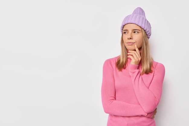 Une jolie adolescente réfléchie aux cheveux blonds considère que quelque chose garde la main sur le menton a une expression pensive douteuse porte un chapeau et un col roulé rose isolé sur blanc avec espace de copie