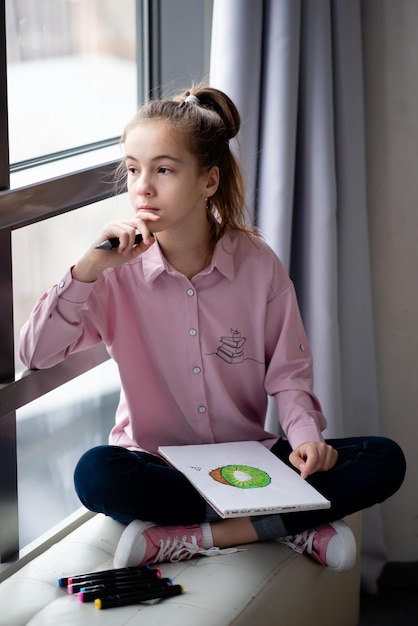 Une jolie adolescente vêtue d'une chemise rose est assise près d'une fenêtre et dessine avec des marqueurs Création