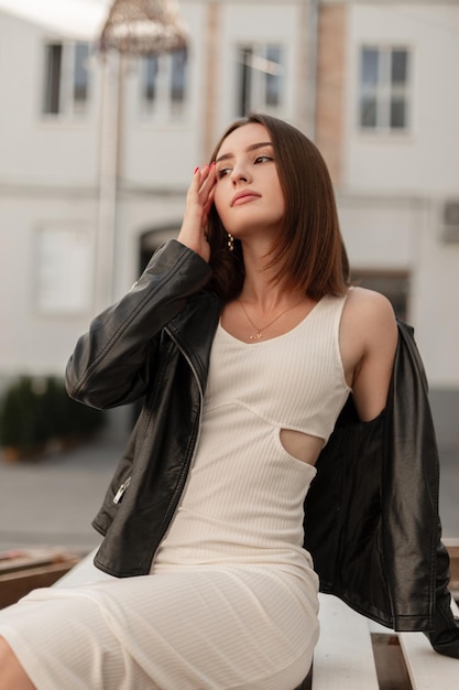 Jolie belle femme caucasienne modèle dans une veste en cuir noir à la mode et une élégante robe blanche s'assoit et se détend dans la ville