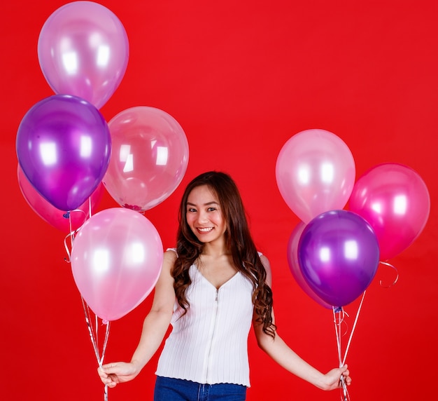 Jolie et belle fille asiatique aux longs cheveux noirs tenant et jouant avec des ballons à air colorés avec un sourire drôle et heureux sur fond rouge, prise de vue en studio.