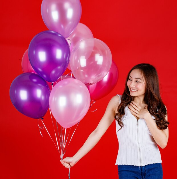 Jolie et belle fille asiatique aux longs cheveux noirs tenant et jouant avec des ballons à air colorés avec un sourire drôle et heureux sur fond rouge, prise de vue en studio.