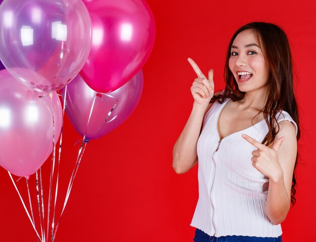 Jolie et belle fille asiatique debout et jouant avec des ballons à air colorés avec un sourire drôle et heureux sur fond rouge, prise de vue en studio. Festival et célébrer le concept.