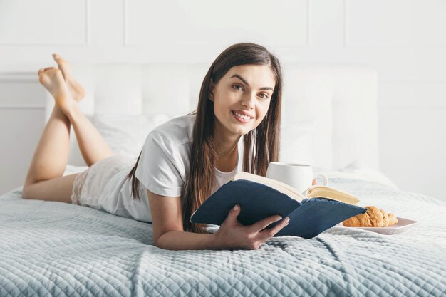 Jolie belle jeune femme lisant un livre et buvant du café sur le lit