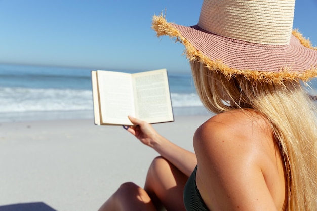 Photo une jolie blonde caucasienne qui passe du temps à la plage par une journée ensoleillée, portant un chapeau de soleil, assise sur une serviette et lisant, avec le ciel bleu et la mer en arrière-plan. vacances estivales sur une plage tropicale.