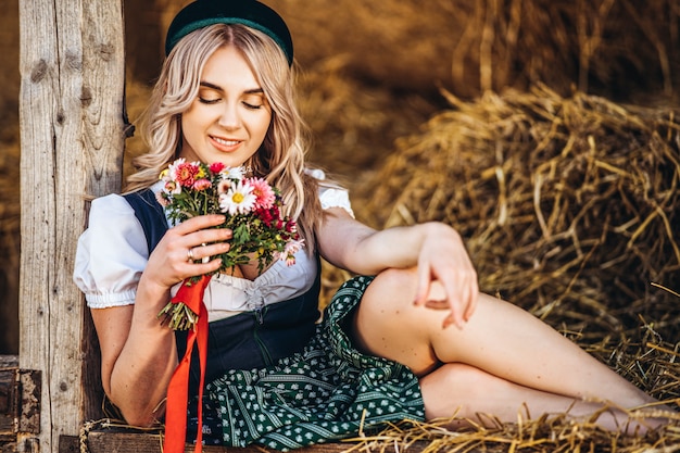 Jolie blonde en dirndl, robe de fête traditionnelle avec bouquet de fleurs des champs assis sur la clôture en bois à la ferme