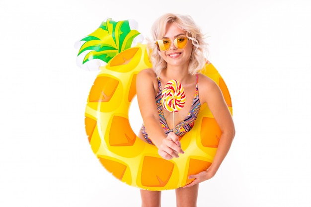 Jolie blonde femme caucasienne se tient en maillot de bain avec anneau en caoutchouc plage ananas et mange une sucette isolé sur blanc