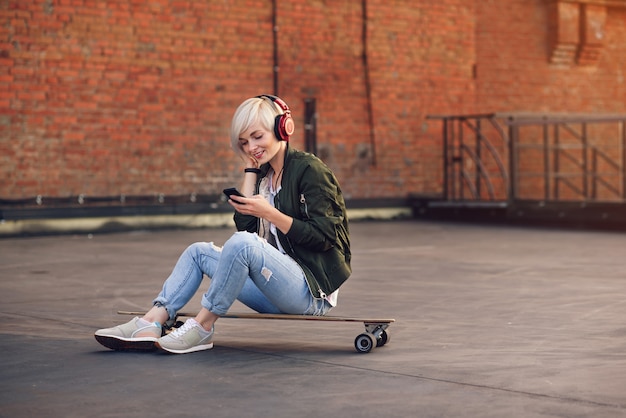 Jolie blonde femme écoutant de la musique dans les écouteurs rouges et à l'aide de téléphone tout en étant assis sur le longboard au mur de briques.