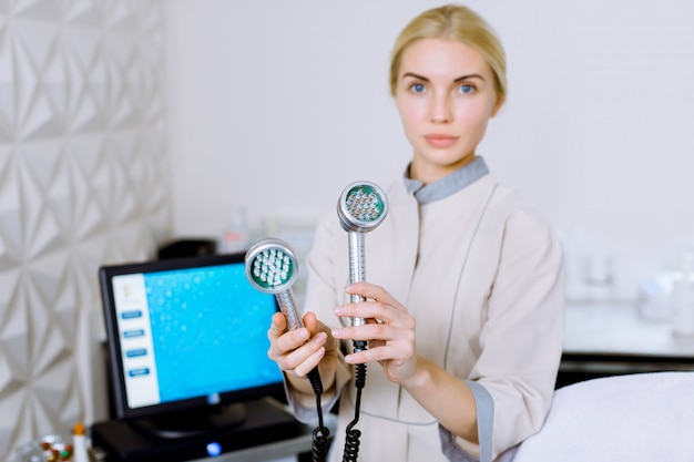 Jolie blonde femme médecin cosmetolotgist et esthéticienne tenant un outil pour la mésothérapie LED Photon Light Therapy RF Rajeunissement de la peau, debout dans un salon de beauté ou une clinique.