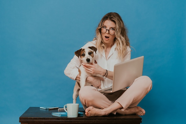 Une jolie blonde à lunettes, vêtue d'une chemise blanche et d'un pantalon rose clair, assise sur une table avec un ordinateur portable, serrant son chiot Jack Russell, semble choquée par la nouvelle avec les yeux et la bouche grands ouverts.