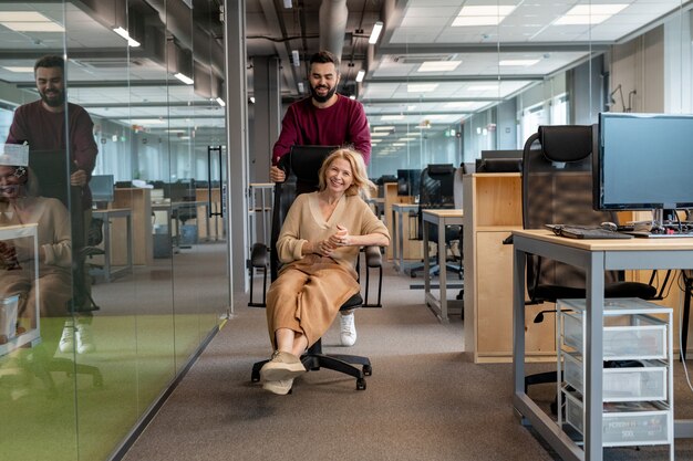 Jolie blonde mature femme d'affaires s'amusant assis dans un fauteuil et son jeune collègue en le poussant le long de l'allée du bureau moderne